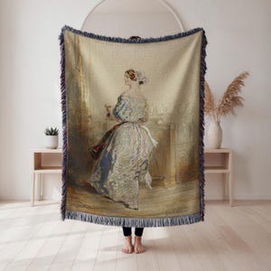 May include: A woven throw blanket featuring a classical painting of a woman in a long dress. The artwork is in muted tones of beige, blue, and red. The blanket has a fringed edge and is displayed in a room with light-colored wood floors and a white wall.