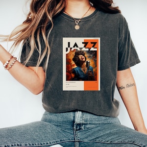 Puede incluir: Una camiseta gris oscuro con un gráfico de estilo vintage de Ella Fitzgerald cantando jazz. El gráfico presenta un retrato de Ella Fitzgerald con la palabra "JAZZ" en letras negritas sobre ella. El gráfico está sobre un fondo naranja y amarillo.