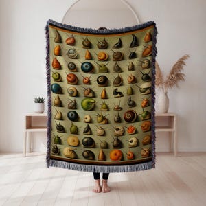 May include: A woven throw blanket featuring a collection of illustrated snails in various shapes and colors against a muted green background. The blanket has a fringed edge and a dark brown border.