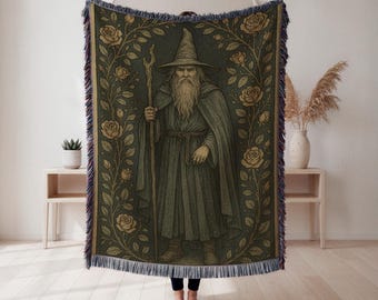 Op de afbeelding: Een geweven sprei met een gedetailleerde illustratie van een tovenaar met staf, omgeven door een bloemenrand van rozen. Het kleurenpalet omvat groen, goud en bruin, met een gefranjerde rand.