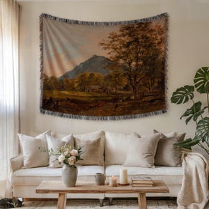 Puede incluir: Un tapiz tejido con una pintura de paisaje de una montaña, árboles y un edificio. El tapiz rectangular tiene un borde con flecos y está colgado sobre un sofá blanco con cojines de colores neutros. Una mesa de centro de madera y objetos decorativos completan la escena.