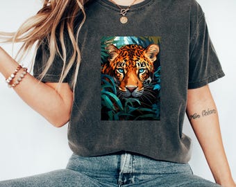 May include: Dark grey t-shirt featuring a vibrant illustration of a cheetah's face with striking blue eyes, surrounded by lush green foliage. The word "Cheetah" is visible in the upper left corner of the image. The shirt is made of a soft, comfortable material.