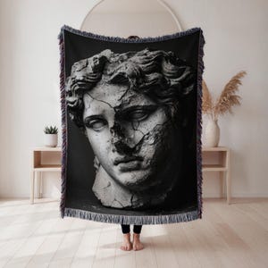 May include: Black and white woven throw blanket featuring a detailed image of a classical statue head. The statue is cracked, creating a dramatic effect. The blanket has a fringed edge with a mix of colors, adding a decorative touch to the home decor item.