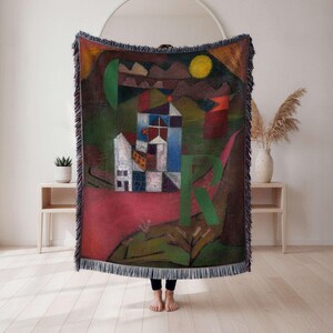 May include: A woven throw blanket featuring a colorful abstract landscape painting. The artwork includes geometric shapes, a house, a yellow sun, and the letter "R". The blanket has a fringed edge.