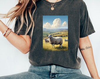May include: A dark grey t-shirt featuring a graphic print of a white sheep in a field with a blue sky and white clouds. The sheep has large horns and is standing in a field of green grass.
