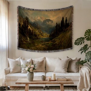 Puede incluir: Un tapiz tejido con una pintura de paisaje de una cordillera, un lago y un bosque. El tapiz tiene un borde con flecos y se muestra sobre un sofá blanco con cojines beige. La paleta de colores incluye verdes, marrones y azules.