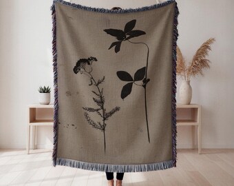 May include: A beige woven throw blanket featuring black botanical illustrations of flowers and leaves. The blanket has a fringed edge with blue, red, and purple accents. The throw is displayed indoors.