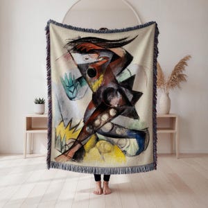 May include: A woven throw blanket featuring an abstract artwork in shades of brown, black, yellow, blue, and gray. The artwork depicts a dynamic figure with geometric shapes and brushstrokes. The blanket has a fringed edge.
