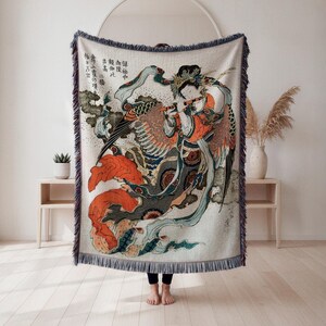 Op de afbeelding: Een geweven sprei met een Japanse houtsnede. Het kunstwerk toont een figuur die fluit speelt, met levendige kleuren zoals oranje, blauw en groen. De sprei heeft een gefranjerde rand en wordt binnenshuis tentoongesteld.