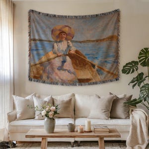 May include: A woven tapestry depicting a painting of a woman in a boat, wearing a large hat. The artwork uses a soft palette of blues, pinks, and yellows. The tapestry is hung above a white sofa with neutral-toned cushions.