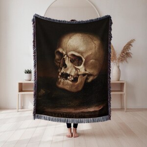 May include: A woven throw blanket featuring a detailed, realistic depiction of a human skull. The skull is rendered in shades of brown and beige against a dark background. The blanket has a fringed edge and the text "TV FVLEGO ERIS" at the bottom.