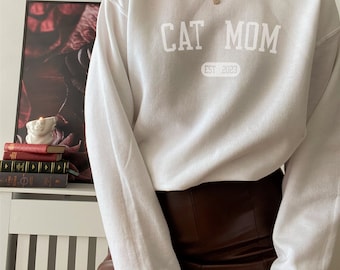 May include: White sweatshirt with the text "CAT MOM EST 2023" in a collegiate font.