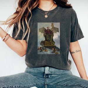 Puede incluir: Una camiseta gris oscuro con una ilustración de estilo vintage de una figura con alas sentada en un trono. La figura sostiene un bastón y está rodeada de figuras más pequeñas.