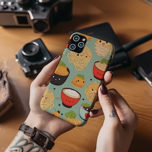 Puede incluir: Una funda para teléfono con un fondo azul claro y un patrón de cuencos de ramen de dibujos animados, fideos y albóndigas. La funda tiene un recorte para el objetivo de la cámara.