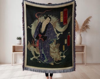 Puede incluir: Una manta verde oscuro con una ilustración de estilo japonés de una figura y un dragón. La figura lleva un kimono azul estampado con una faja roja. La manta tiene un borde con flecos.
