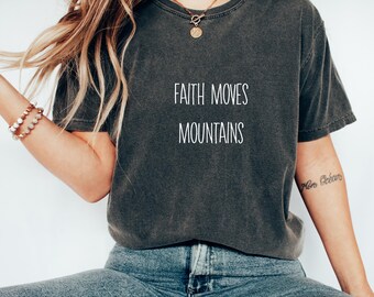 Puede incluir: Una camiseta gris oscuro con el texto "FAITH MOVES MOUNTAINS" impreso en blanco en la parte delantera. La camiseta la lleva puesta una persona con el pelo largo y castaño.