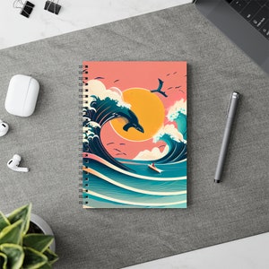 May include: A spiral bound notebook with a colorful illustration of a whale jumping over a large wave with a bright yellow sun in the background. The wave is blue and green with white foam. The background is a light pink color.