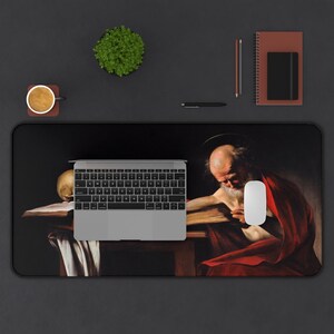 May include: A black and white desk mat with a painting of a man in red robes reading a book. The man is sitting at a table with a skull on it. A laptop computer and a white mouse are on the desk mat.