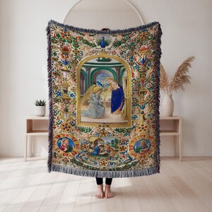 Könnte beinhalten: Eine gewebte Überwurfdecke mit einem religiösen Gemälde im Renaissance-Stil. Das zentrale Bild zeigt einen Engel und eine Frau in blauen Gewändern. Die Decke hat einen dekorativen Rand mit floralen und verzierten Details und einen Fransenrand.