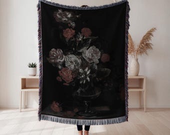May include: Black woven throw blanket with a floral still life design. The artwork shows a bouquet of pink and white flowers in a glass vase against a dark backdrop. The blanket has a fringed edge.