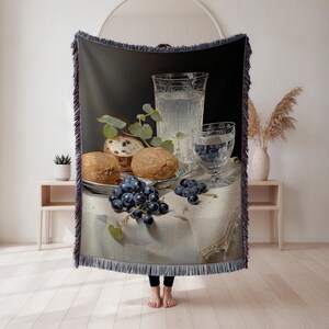 May include: A woven throw blanket featuring a still life painting with bread rolls, grapes, and glasses of water. The artwork has a vintage aesthetic with a dark background and a white tablecloth. The blanket has a fringed edge.