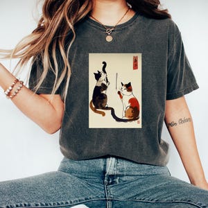 May include: A dark gray t-shirt with a vintage-style illustration of two cats playing with a string. The illustration is in black, white, and red on a cream background. The text "猫" is visible in the upper right corner of the illustration.