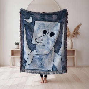 May include: A blue and white woven throw blanket featuring an abstract figure with a crescent moon. The artwork has a whimsical style with black outlines and a fringe border. The blanket is displayed indoors.