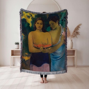 May include: A woven throw blanket featuring a painting of two women holding a watermelon. The artwork has a green, yellow, and blue color palette with a fringe border. The blanket is displayed in a room with a white wall and wooden floor.