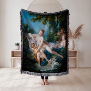 May include: Woven throw blanket featuring a classical painting with figures in a forest setting. The artwork depicts a woman with two cherubs and doves, set against a backdrop of lush greenery and a blue sky. The blanket has a fringed edge.