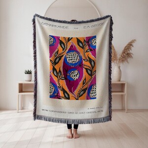 Könnte beinhalten: Eine beige Decke mit einem farbenfrohen zentralen Design. Das Design zeigt stilisierte Blumen in Orange, Lila und Blau mit schwarzen Blättern. Die Decke hat einen Fransenrand und Text oben und unten.