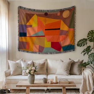 May include: A colorful woven wall tapestry featuring an abstract geometric design in shades of orange, yellow, red, and blue. The tapestry is rectangular with a fringed edge and is displayed above a white sofa in a living room setting.