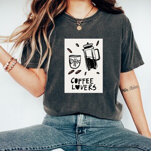 May include: A dark grey t-shirt with a white graphic of a coffee cup and a French press. The graphic also includes the text "Coffee Lovers" with a heart symbol.