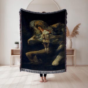 Puede incluir: Una manta tejida negra con una reproducción de la pintura de Goya "Saturno devorando a su hijo". La obra de arte representa una figura monstruosa que consume a una figura más pequeña, con colores oscuros y apagados. La manta tiene un borde con flecos.