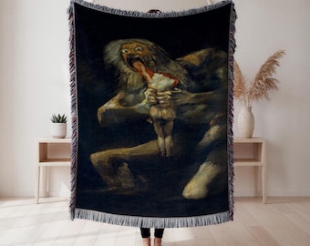Può includere: Una coperta nera tessuta con una riproduzione del dipinto di Goya "Saturno che divora suo figlio". L'opera raffigura una figura mostruosa che consuma una figura più piccola, con colori scuri e tenui. La coperta ha un bordo sfrangiato.