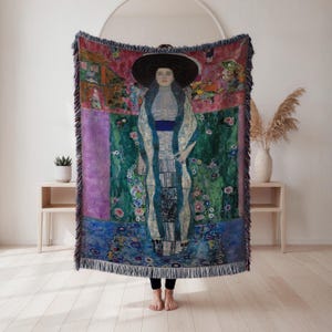 Puede incluir: Una manta tejida con una reproducción de una pintura de Gustav Klimt. La obra de arte representa a una mujer con un vestido largo y un sombrero grande, sobre un fondo de elementos florales y paisajísticos coloridos. La manta tiene bordes con flecos.