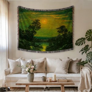 May include: A woven tapestry featuring a landscape painting with a sunset over a lake. The artwork is predominantly green and yellow, with trees and water in the foreground. The tapestry is hanging above a white sofa with beige pillows.