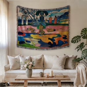 May include: A woven tapestry depicting a colorful scene with figures, trees, and water. The artwork features a variety of colors, including blue, pink, yellow, and green. The tapestry is rectangular with a fringed edge and is hanging on a wall above a white sofa.