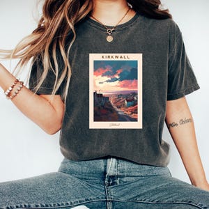 May include: A dark gray t-shirt with a graphic print of a sunset over a town with the text "KIRKWALL, Shetland" on a light peach background.