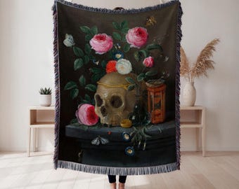 May include: A woven throw blanket with a still life painting. The artwork depicts a skull, roses, and butterflies against a dark background, with a fringed border. The colour scheme includes pink, green, and gold.