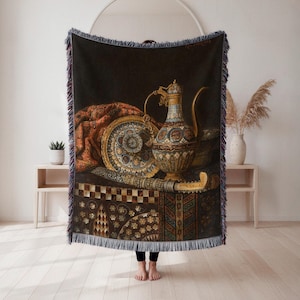 May include: A woven throw blanket featuring a still life painting with a decorative plate, ornate pitcher, and a decorative knife. The blanket has a dark background with a fringed edge. The color palette includes gold, brown, and red.