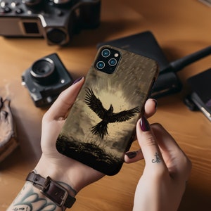 May include: A phone case featuring a black raven with outstretched wings against a sepia-toned background. The case has a matte finish and is held in a person's hands. The phone case is designed to fit a smartphone.