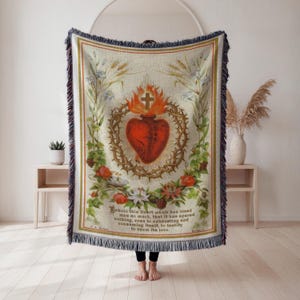 May include: Cream-colored throw blanket featuring a central illustration of a red heart encircled by a crown of thorns, a cross, and flames. The design is surrounded by floral and botanical elements, with text at the bottom. The blanket has a fringed edge.