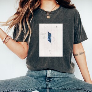 Puede incluir: Una camiseta gris oscuro con un estampado gráfico blanco de una ilustración azul y blanca. La ilustración es un diseño geométrico con el texto "FLORA NOTA" y "34" impreso encima.