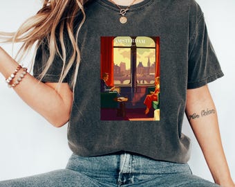 May include: A dark gray t-shirt with a vintage-style graphic of a couple sitting by a window overlooking the city of Amsterdam. The graphic has the text "AMSTERDAM" at the top.