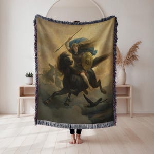 May include: Woven throw blanket with a classical painting of a warrior on horseback. The artwork shows a woman with a spear and shield, riding a black horse. The blanket has a fringed edge and is displayed against a neutral background.