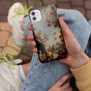 Puede incluir: Una funda de tel&eacute;fono blanca con una pintura colorida de estilo renacentista de &aacute;ngeles y figuras. La funda tiene una protuberancia de c&aacute;mara elevada con tres lentes.
