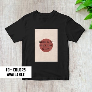 May include: Black t-shirt with a graphic print of a red circle with the text "THERE IS NO PLACE LIKE 127.0.0.1". The text "10+ COLORS AVAILABLE" is printed on a white rectangle below the shirt.