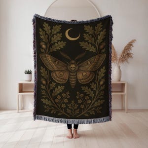 May include: Black throw blanket with a large moth design, a crescent moon, and botanical details. The moth is brown and tan, with green and gold accents. The blanket has a fringed edge.