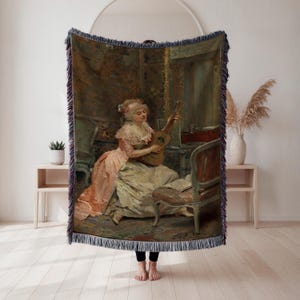 May include: A woven throw blanket featuring a classical painting of a woman playing a lute. The artwork depicts a woman in a pink and cream dress, seated in a room with antique furniture. The blanket has a fringed edge and is displayed against a white wall.
