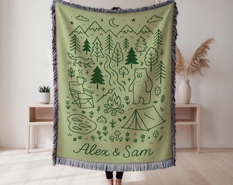 May include: A green throw blanket with a camping scene design in dark green. The design includes trees, mountains, a fox, a bear, a campfire, and a tent. The names "Alex & Sam" are printed at the bottom. The blanket has a fringed edge.